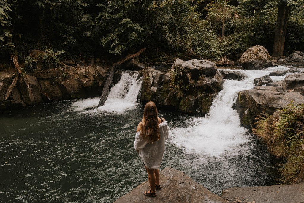 a woman with long hair and white top stands before a small waterfall and pool in La Fortuna free things to do
