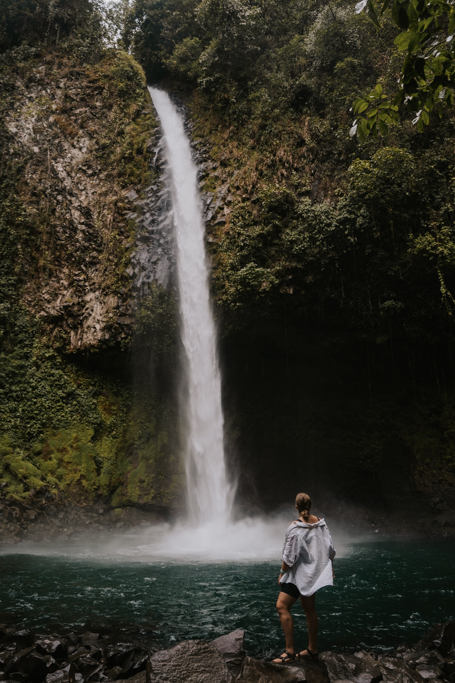 a woman in a white shirt stands in front of La Fortuna Waterfall, one of the best La Fortuna things to do