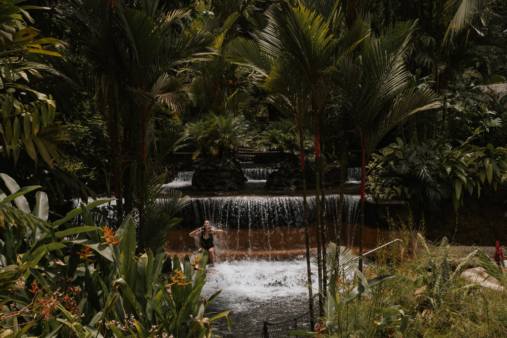 a woman in a black swimming suit sits on the edge of a tiered hot spring at Tabacon, one of the best things to do near La Fortuna