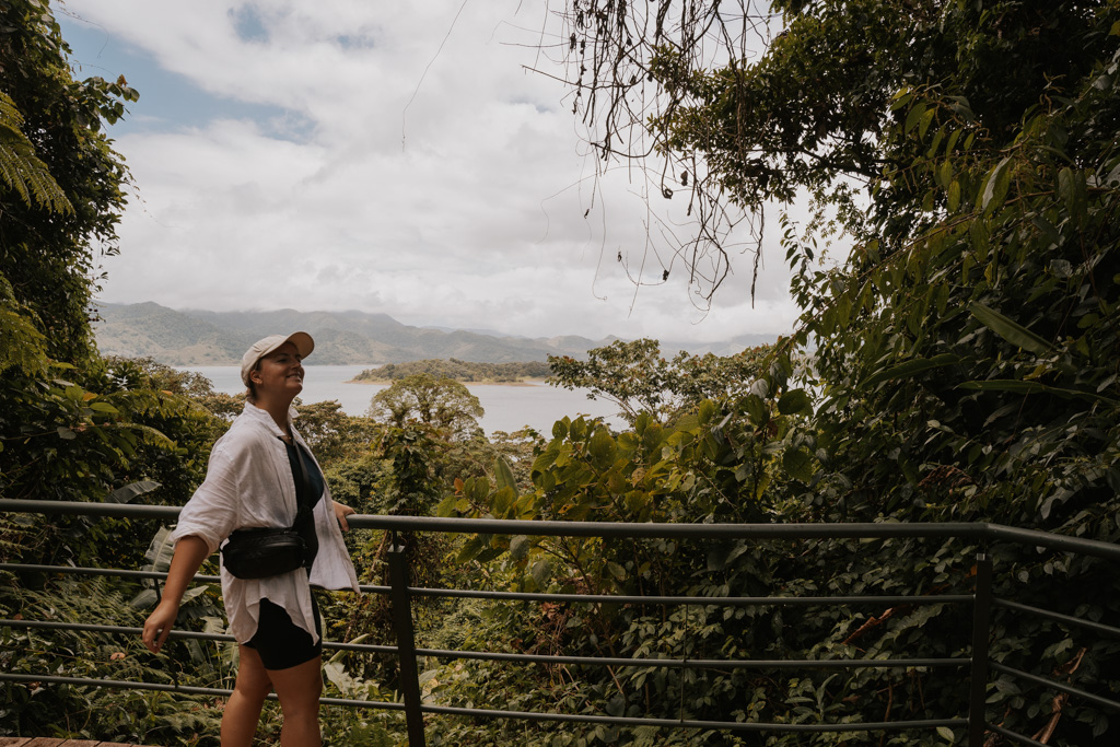 a woman smiles wearing a white cap and shirt with black purse in front of a jungle with Lake Arenal La Fortuna Costa Rica in the distance