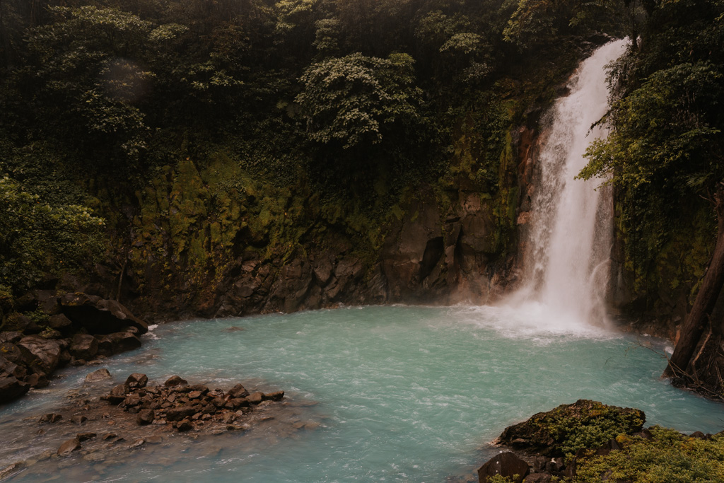 Rio Celeste Waterfall gushes into an electric green blue pool below with rugged cliffs, one of the best day trips from La Fortuna Costa Rica