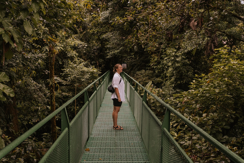 a woman in a white shirt and black shorts walks on a green suspension bridge amongst Costa Rica rainforest