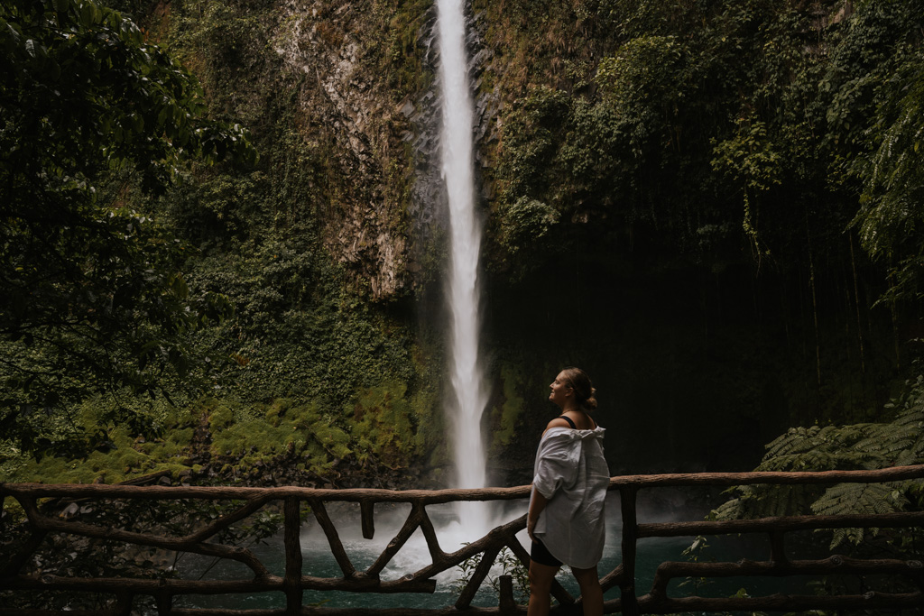La Fortuna Waterfall in Costa Rica stands behind a girl in a loose white shirt