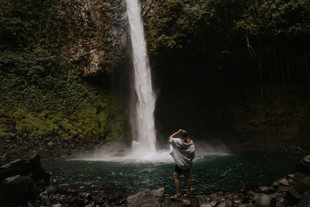 a woman in a white shirt stands before the thundering La Fortuna Waterfall in Costa Rica with lush dark green surrounds