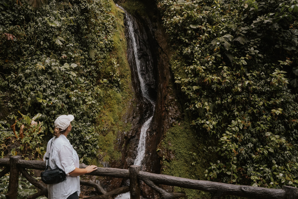 a woman in a white hat and shirt looks up at a waterfall in hanging bridges arenal costa rica