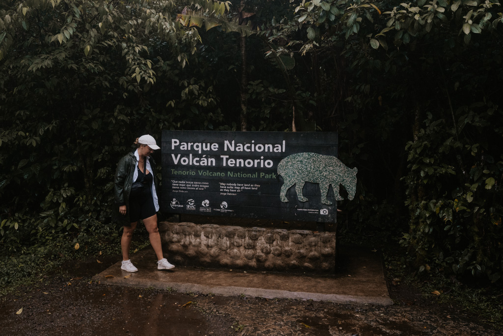 a woman with white hat and sneakers stands in front of a dark wood sign that reads 'parque nacional volcan tenorio'