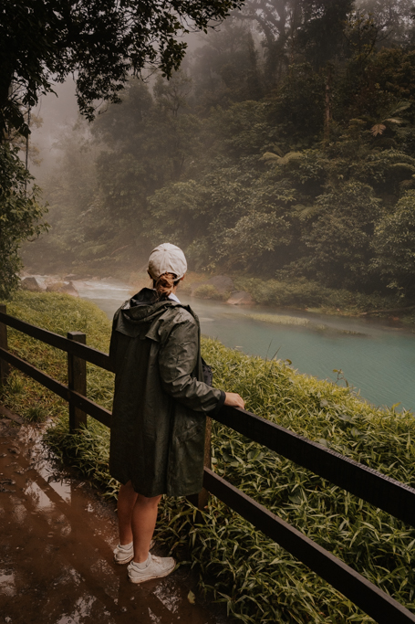 a woman in green rain jacket and white hat stands at a railing looking over the stream leading to rio celeste waterfalls