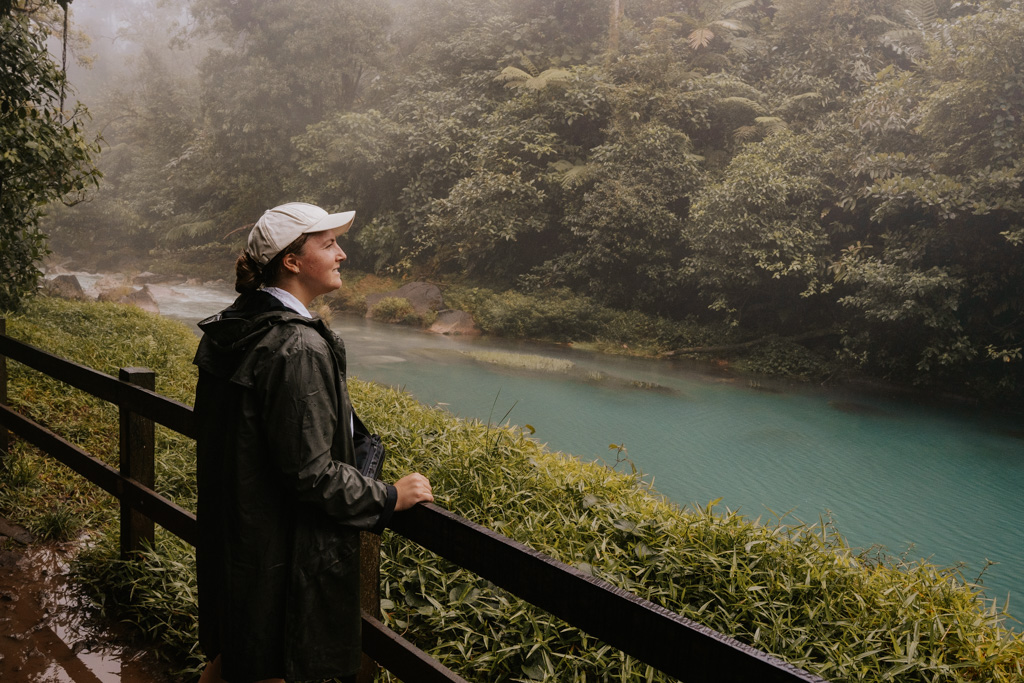 Haley Blackall wears a green rain jacket and white hat standing in front of the river celeste costa rica