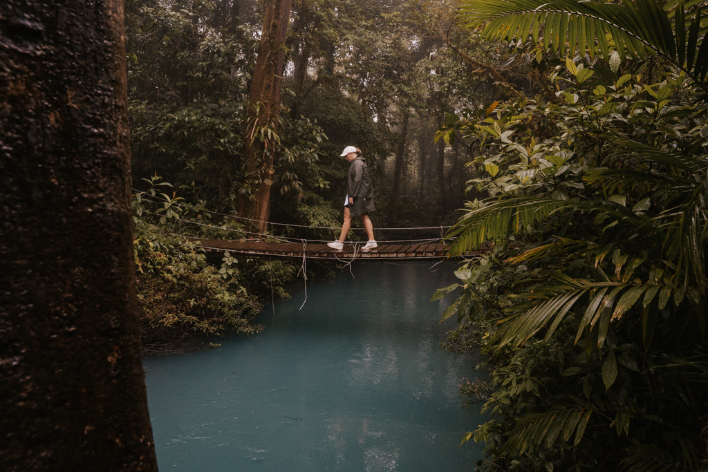 a woman in white hat walks along a wooden bridge over blue water and dense jungle