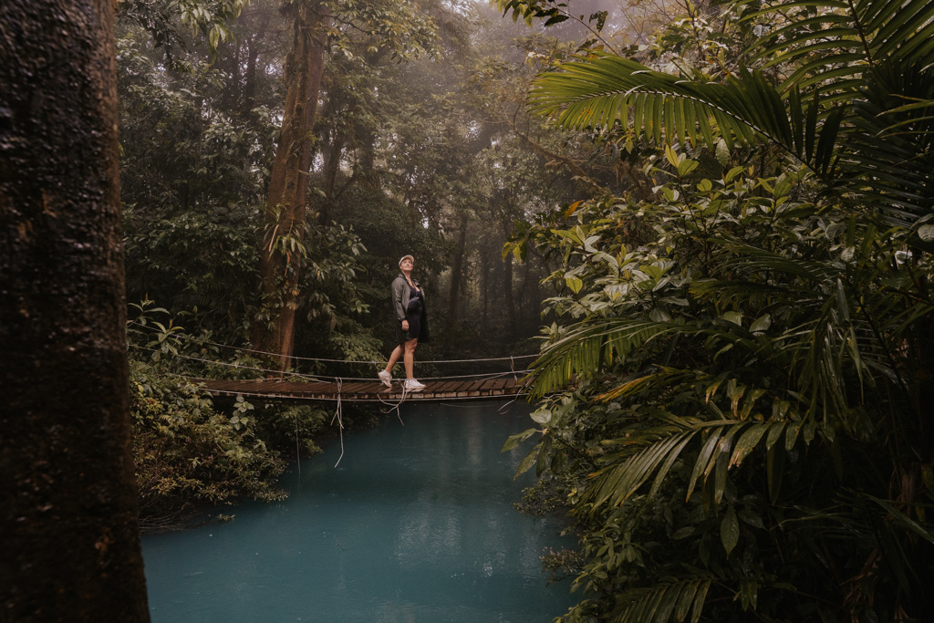 a woman in white sneakers walks across a wooden bridge about the river at costa rica rio celeste
