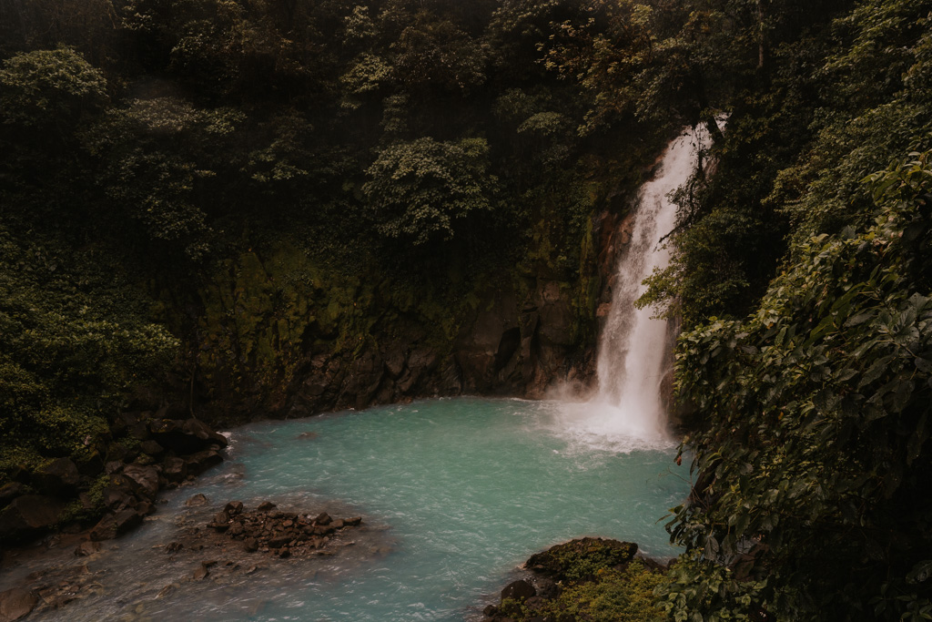 tour rio celeste waterfall with gushing waters over a blue pond and dark green jungle