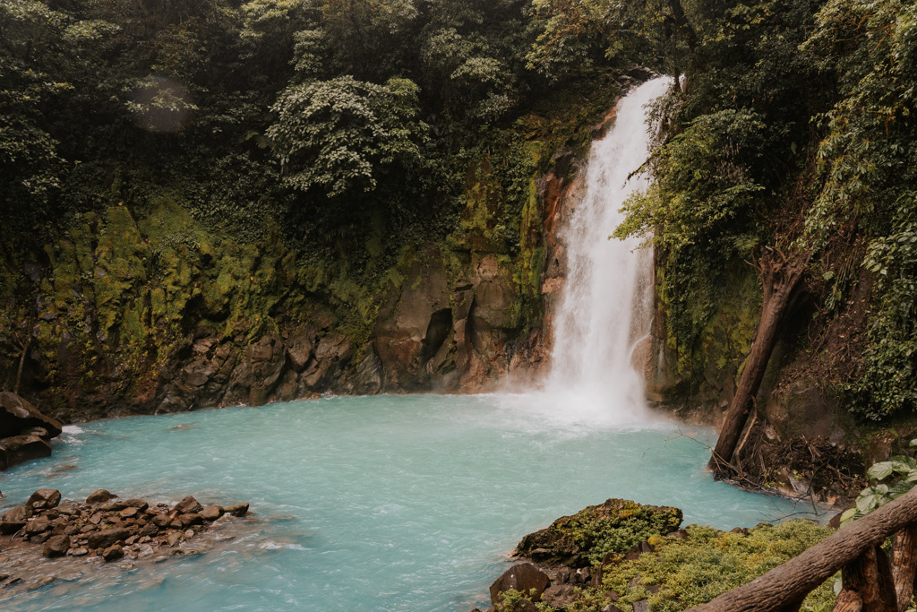 electric blue waters sit below a roaring rio celeste waterfall amongst dark green rainforest